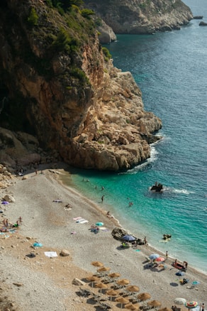 A beautiful rocky coastline with turquoise waters where people are enjoying sunbathing and swimming. The beach is lined with pebbles, and there are sun umbrellas and beach towels scattered along the shore. The landscape is dominated by steep cliffs with lush greenery on top, adding to the natural beauty of the setting.