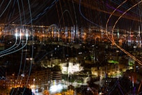A vibrant nighttime cityscape with a striking display of light trails and illuminated buildings. The scene is filled with colorful streaks of light across the sky, possibly from long exposure photography. The city lights twinkle in the distance, casting a warm glow over the urban environment.