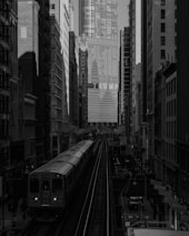 A busy urban scene featuring a train traveling on elevated tracks between tall buildings. The background prominently displays a high-rise building with the word 'TRUMP' in large letters. The environment appears densely built, with various structures tightly packed on either side of the tracks. The image is presented in black and white, adding a dramatic and slightly somber tone to the cityscape.
