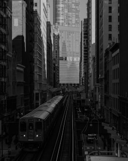 A busy urban scene featuring a train traveling on elevated tracks between tall buildings. The background prominently displays a high-rise building with the word 'TRUMP' in large letters. The environment appears densely built, with various structures tightly packed on either side of the tracks. The image is presented in black and white, adding a dramatic and slightly somber tone to the cityscape.