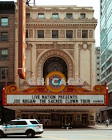 An ornate theater entrance with the word 'CHICAGO' prominently displayed on a marquee. Below it, there's an announcement for a tour titled 'Joe Rogan: The Sacred Clown Tour.' A police vehicle marked 'Chicago Police Transit Detail' is parked in front of the theater. The building features detailed architectural elements including arches and intricate stonework. The area is surrounded by other buildings, including a modern glass structure to the right.
