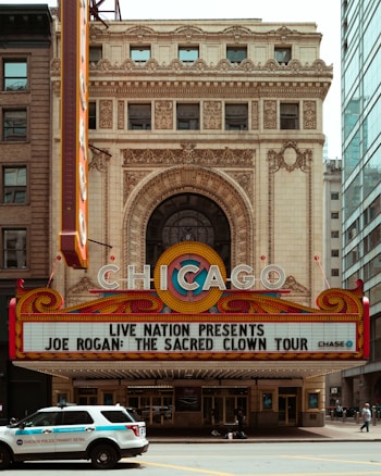 An ornate theater entrance with the word 'CHICAGO' prominently displayed on a marquee. Below it, there's an announcement for a tour titled 'Joe Rogan: The Sacred Clown Tour.' A police vehicle marked 'Chicago Police Transit Detail' is parked in front of the theater. The building features detailed architectural elements including arches and intricate stonework. The area is surrounded by other buildings, including a modern glass structure to the right.