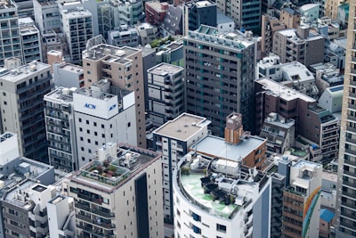 A dense urban environment features numerous high-rise buildings packed closely together. The architecture varies from modern glass structures to older brick and concrete buildings, with many having rooftop equipment such as air conditioning units and antennas. A mix of flat and slightly inclined rooftops can be observed, with some showing greenery or satellite dishes.