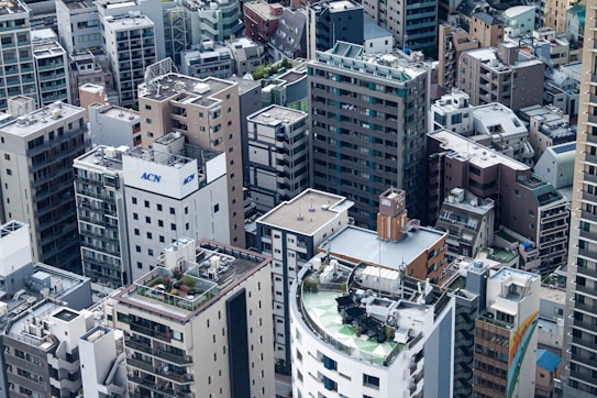 A dense urban environment features numerous high-rise buildings packed closely together. The architecture varies from modern glass structures to older brick and concrete buildings, with many having rooftop equipment such as air conditioning units and antennas. A mix of flat and slightly inclined rooftops can be observed, with some showing greenery or satellite dishes.