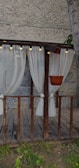 A wooden pergola decorated with sheer white curtains tied to its pillars. The structure includes a hanging potted plant, and is surrounded by a gravel-textured wall. Small string lights are attached to the top beam of the pergola, creating an inviting atmosphere.