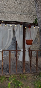 A wooden pergola decorated with sheer white curtains tied to its pillars. The structure includes a hanging potted plant, and is surrounded by a gravel-textured wall. Small string lights are attached to the top beam of the pergola, creating an inviting atmosphere.