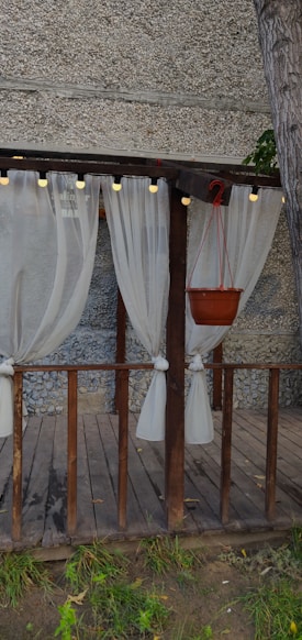 A wooden pergola decorated with sheer white curtains tied to its pillars. The structure includes a hanging potted plant, and is surrounded by a gravel-textured wall. Small string lights are attached to the top beam of the pergola, creating an inviting atmosphere.