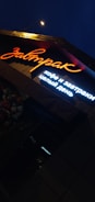 A brightly lit sign in Russian characters is shown under night lighting, with the main word illuminated in orange neon and additional text below in white light. Adjacent to the sign, there is a cluster of flowers visible, enhancing the ambient outdoor setting.