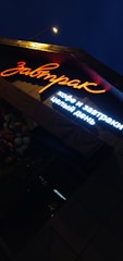 A brightly lit sign in Russian characters is shown under night lighting, with the main word illuminated in orange neon and additional text below in white light. Adjacent to the sign, there is a cluster of flowers visible, enhancing the ambient outdoor setting.
