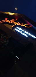 A brightly lit sign in Russian characters is shown under night lighting, with the main word illuminated in orange neon and additional text below in white light. Adjacent to the sign, there is a cluster of flowers visible, enhancing the ambient outdoor setting.