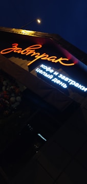 A brightly lit sign in Russian characters is shown under night lighting, with the main word illuminated in orange neon and additional text below in white light. Adjacent to the sign, there is a cluster of flowers visible, enhancing the ambient outdoor setting.