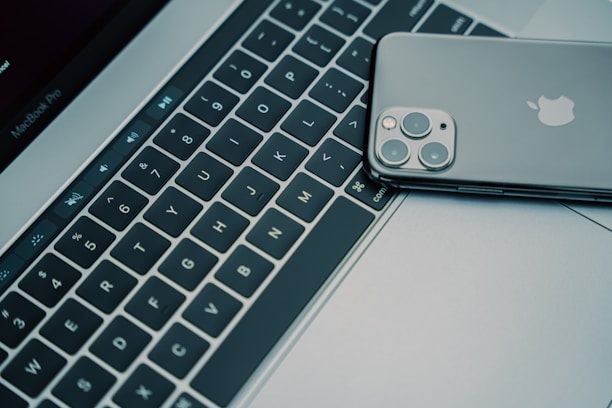 A close-up view of a laptop keyboard with an Apple laptop partially visible in the background. A smartphone with a reflective back, displaying the Apple logo and cameras, is placed on the keyboard.