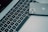 A close-up view of a laptop keyboard with an Apple laptop partially visible in the background. A smartphone with a reflective back, displaying the Apple logo and cameras, is placed on the keyboard.
