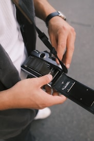 Hands are handling a film camera, adjusting or loading film into it. The camera is open, revealing the film chamber, and the person's attire includes a watch and casual clothing.