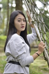 A young woman with long dark hair stands outdoors, holding onto a net-like structure. She is wearing a light gray sweater and a black belt, and the background features blurred green foliage, indicating a natural setting.