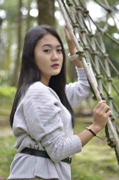 A young woman with long dark hair stands outdoors, holding onto a net-like structure. She is wearing a light gray sweater and a black belt, and the background features blurred green foliage, indicating a natural setting.