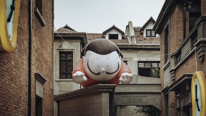 A large cartoon inflatable figure with a smiling face is suspended between two brick buildings. The figure is painted with a black hairstyle and red outfit, appearing to lean forward with hands outstretched. The surrounding architecture includes traditional brickwork and tiled rooftops, typical of older urban environments.