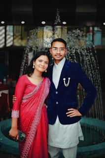 A couple stands in front of an elegant water fountain. The woman is wearing a red traditional dress with intricate lace patterns and is holding a smartphone. The man is dressed in a navy blue velvet blazer with a white traditional kurta underneath, featuring a decorative chain. Both appear to be smiling softly, suggesting a formal or celebratory occasion.