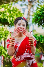 A woman dressed in a traditional red sari adorned with white embroidery. She is smiling and displaying her hands, which are decorated with henna. Gold jewelry, including bracelets and a necklace, complements her attire. The background is lush with greenery, enhancing the vibrant scene.