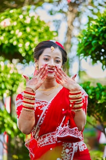 A woman dressed in a traditional red sari adorned with white embroidery. She is smiling and displaying her hands, which are decorated with henna. Gold jewelry, including bracelets and a necklace, complements her attire. The background is lush with greenery, enhancing the vibrant scene.