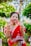 A woman dressed in a traditional red sari adorned with white embroidery. She is smiling and displaying her hands, which are decorated with henna. Gold jewelry, including bracelets and a necklace, complements her attire. The background is lush with greenery, enhancing the vibrant scene.