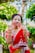 A woman dressed in a traditional red sari adorned with white embroidery. She is smiling and displaying her hands, which are decorated with henna. Gold jewelry, including bracelets and a necklace, complements her attire. The background is lush with greenery, enhancing the vibrant scene.
