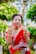 A woman dressed in a traditional red sari adorned with white embroidery. She is smiling and displaying her hands, which are decorated with henna. Gold jewelry, including bracelets and a necklace, complements her attire. The background is lush with greenery, enhancing the vibrant scene.