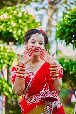 A woman dressed in a traditional red sari adorned with white embroidery. She is smiling and displaying her hands, which are decorated with henna. Gold jewelry, including bracelets and a necklace, complements her attire. The background is lush with greenery, enhancing the vibrant scene.