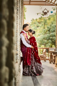 A couple stands closely together in a warm embrace on a stone-lined veranda. The woman wears a richly embroidered maroon sari with intricate patterns, complemented by a matching dupatta. The man is dressed in a maroon waistcoat over a white shirt. Greenery and wooden furniture are visible in the background, alongside a hanging lamp, creating a serene and romantic setting.