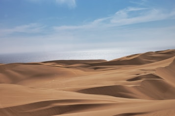 Vast golden sand dunes stretch across the landscape, meeting the horizon where a shimmering body of water lies under a bright blue sky.