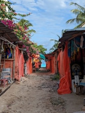 A narrow sandy path is flanked by market stalls covered with orange tarps. Vibrant artwork and handicrafts are visible, including a colorful painting of a face. Pink bougainvillea flowers and palm trees add a tropical vibe. In the distance, the sea is visible under a partly cloudy sky.