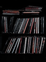 A collection of music CDs is organized on a shelf. The CDs are in thin plastic cases, arranged vertically and horizontally. A variety of titles and artists can be seen on the spines, with different colors and fonts.