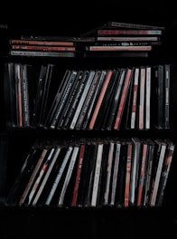 A collection of music CDs is organized on a shelf. The CDs are in thin plastic cases, arranged vertically and horizontally. A variety of titles and artists can be seen on the spines, with different colors and fonts.