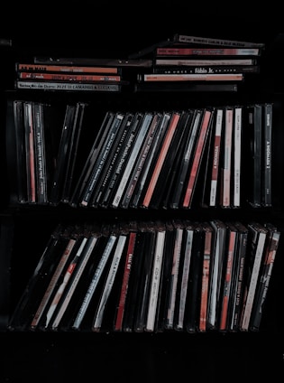 A collection of music CDs is organized on a shelf. The CDs are in thin plastic cases, arranged vertically and horizontally. A variety of titles and artists can be seen on the spines, with different colors and fonts.
