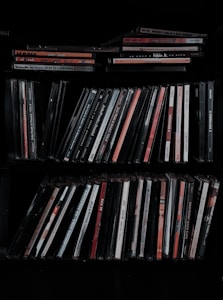A collection of music CDs is organized on a shelf. The CDs are in thin plastic cases, arranged vertically and horizontally. A variety of titles and artists can be seen on the spines, with different colors and fonts.