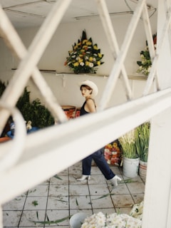 A person wearing a white cowboy hat and dark clothing appears to be in a flower shop. Various floral arrangements are displayed on the walls and floor, with bright colors such as yellows, reds, and greens. The scene is viewed through a white lattice or grid structure, adding an element of depth and framing.