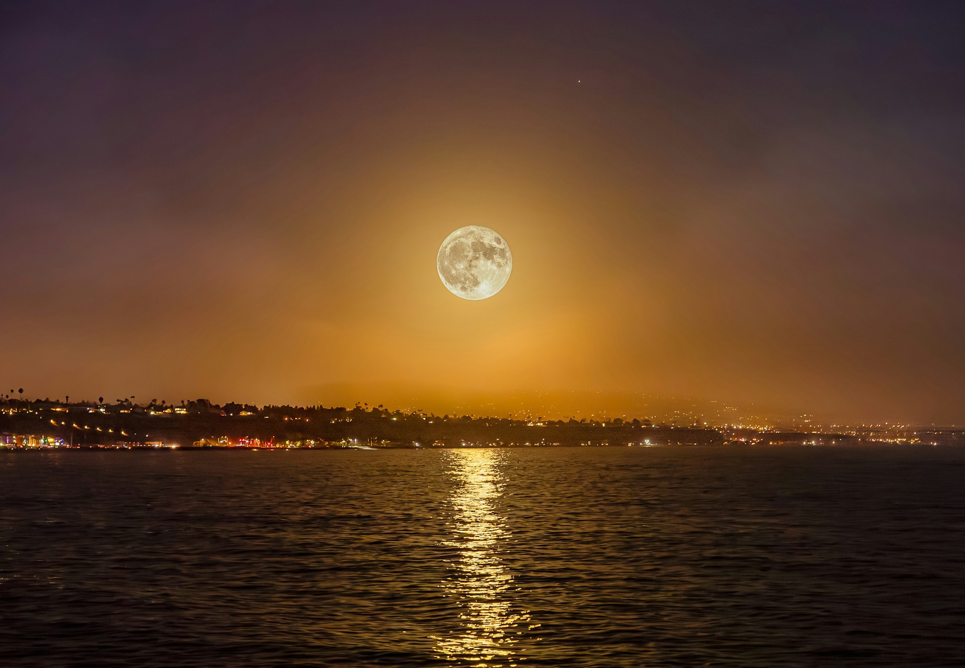a full moon rising over the ocean with a city in the background