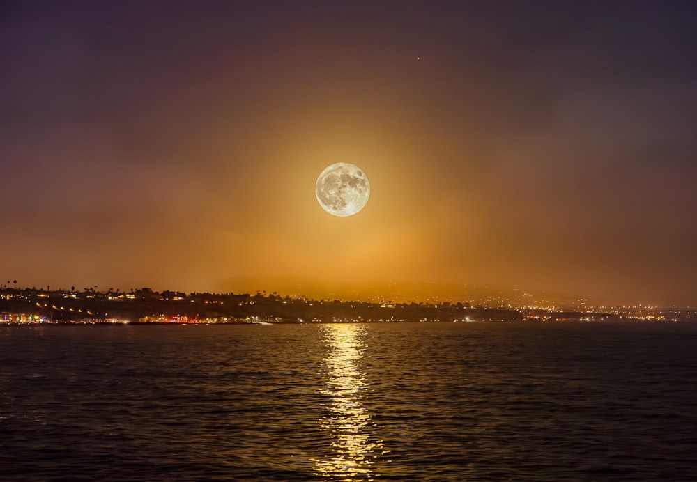 a full moon rising over the ocean with a city in the background