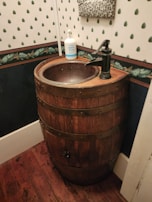 A rustic bathroom sink created from a wooden barrel, featuring a bronze faucet. A bottle of hand soap is placed on the rim. The walls have a vintage style with patterned wallpaper and wood trim, and the floor is made of dark wooden planks.