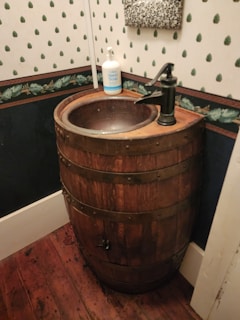A rustic bathroom sink created from a wooden barrel, featuring a bronze faucet. A bottle of hand soap is placed on the rim. The walls have a vintage style with patterned wallpaper and wood trim, and the floor is made of dark wooden planks.