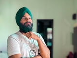 A person with a thoughtful expression stands indoors wearing a green turban and a gray T-shirt with a logo on it. The background is softly blurred, highlighting a doorway and some indistinct objects.