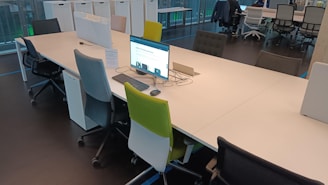A modern office space features a long shared desk with several ergonomic office chairs in various colors, including grey, blue, and green. A computer monitor is placed on one side of the desk, displaying a webpage. The room is well-lit with natural light coming from large windows. In the background, more desks and chairs are visible, along with storage units against the wall.
