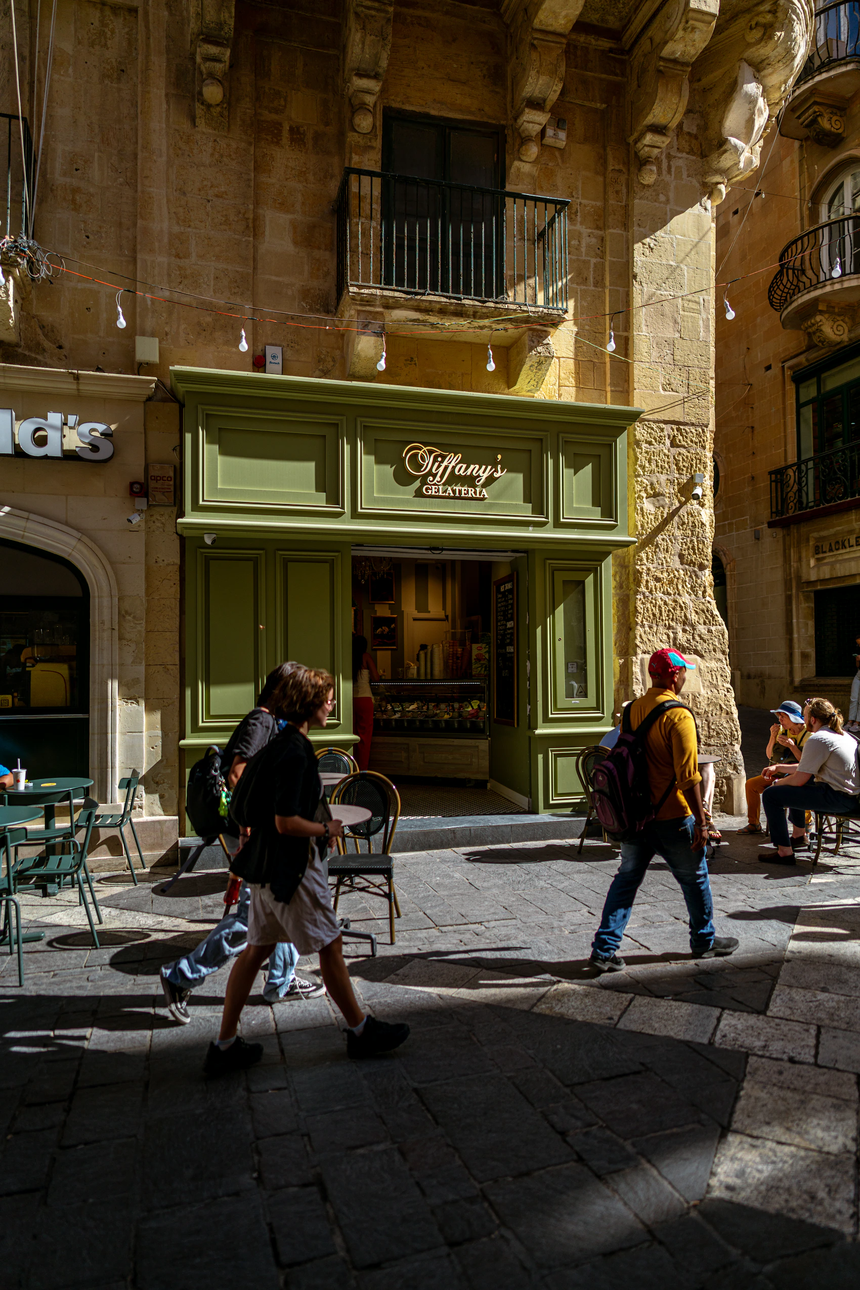 People passing a small green gelateria beneath historic limestone balconies in Valletta
