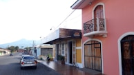 A quiet street scene featuring colorful buildings, one painted in pink with elegant wrought-iron details, and another building adorned in yellow with a sign that reads 'Calidad y Tradición.' A mountain is visible in the background, and a few cars and a motorcycle are parked on the road.