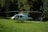 A white helicopter is parked on a grassy field with dense green trees and a wooden cabin in the background. The rotor blades are stationary, and the helicopter bears a logo and registration markings on the side.