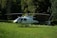A white helicopter is parked on a grassy field with dense green trees and a wooden cabin in the background. The rotor blades are stationary, and the helicopter bears a logo and registration markings on the side.