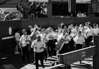 A group of uniformed musicians is marching together, each holding a musical instrument like drums and brass instruments. Behind them, a crowd of photographers is capturing the scene from an elevated position. The atmosphere suggests a formal event or procession. The image is in black and white, adding a timeless feel.