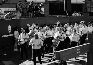 A group of uniformed musicians is marching together, each holding a musical instrument like drums and brass instruments. Behind them, a crowd of photographers is capturing the scene from an elevated position. The atmosphere suggests a formal event or procession. The image is in black and white, adding a timeless feel.