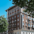 A multi-story brick building with classic architectural details, including ornate balconies and white window frames. The building features a vertical sign reading 'BALKANTOURIST' in blue letters. Surrounding the building, there are leafy green trees, and the sky is clear and blue.