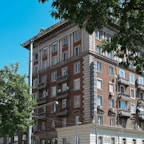 A multi-story brick building with classic architectural details, including ornate balconies and white window frames. The building features a vertical sign reading 'BALKANTOURIST' in blue letters. Surrounding the building, there are leafy green trees, and the sky is clear and blue.